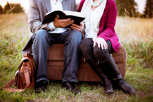 husband and wife seated on a cushioned bench, in an open field with the husband having an open bible in his hands